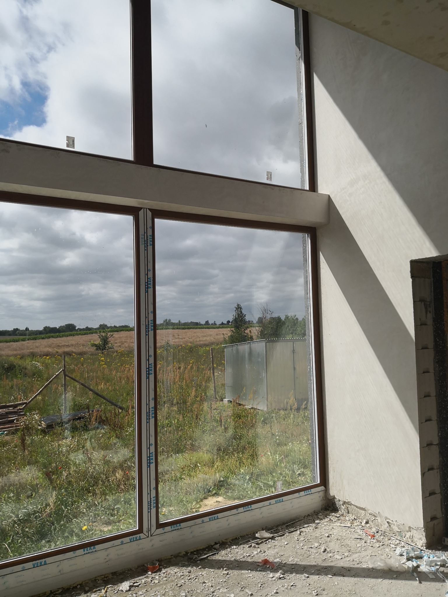 Świeżo położone tynki maszynowe na ścianie wewnętrznej budynku w stanie surowym, widok na pole za oknem z brązową ramą, ślady po pracach wykończeniowych na podłodze.