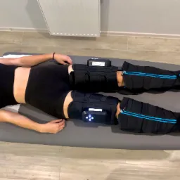 Kobieta leży na leżance z założonymi mankietami pneumatycznego masażu uciskowego na nogach, podłączonymi do urządzenia sterującego.