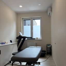Maciej Kwiatkowski - Fizjotywacja - Jasne wnętrze gabinetu z kozetką, bieżnią i biurkiem. Na parapecie modelu czaszki. Minimalistyczny wystrój, stonowana kolorystyka, światło dzienne.