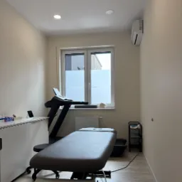 Jasne wnętrze gabinetu z kozetką, bieżnią i biurkiem. Na parapecie modelu czaszki. Minimalistyczny wystrój, stonowana kolorystyka, światło dzienne.