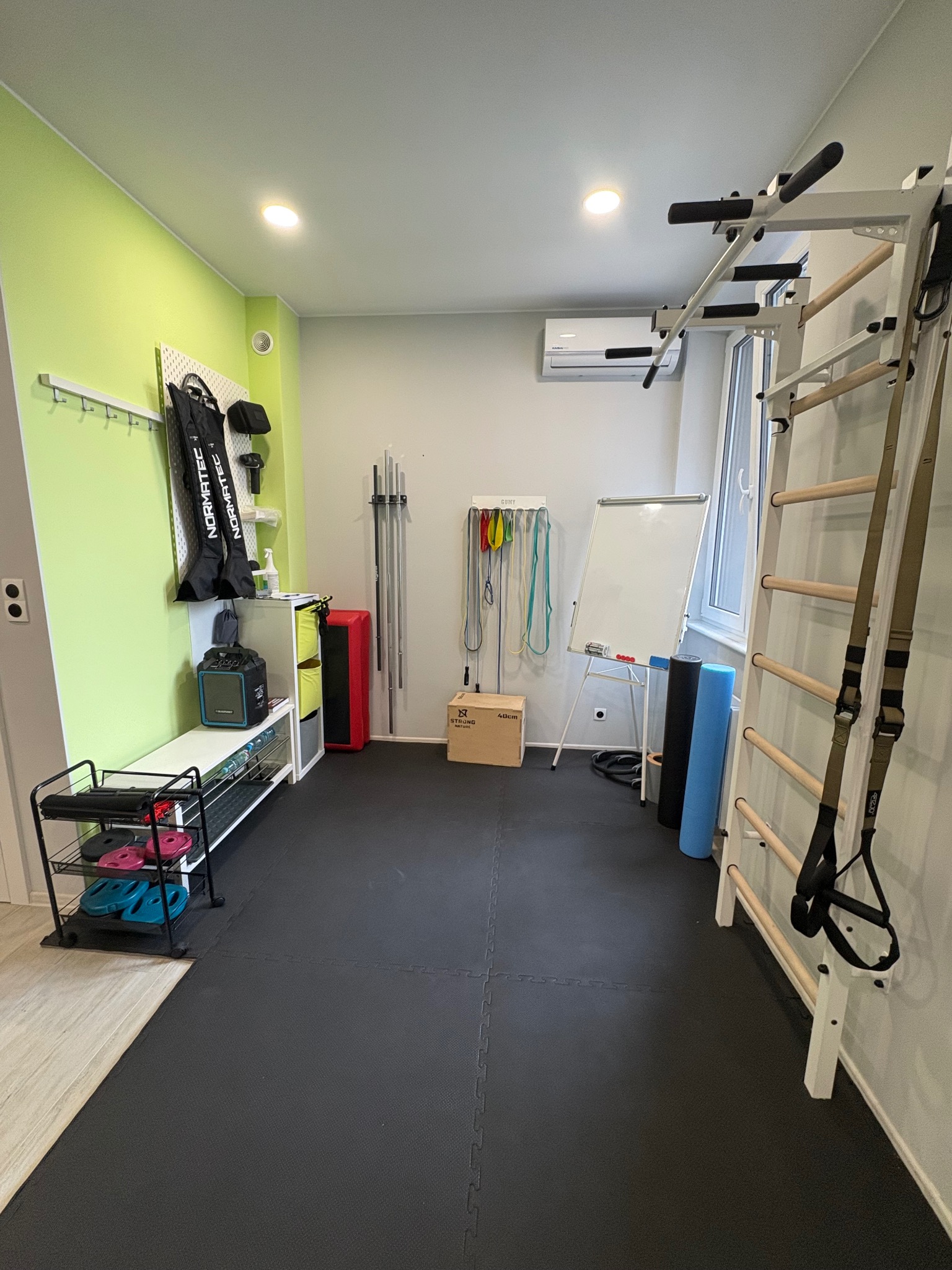 Jasne wnętrze sali do ćwiczeń z drabinką gimnastyczną, gumami oporowymi, stepem i tablicą. Zielona ściana z akcesoriami. Podłoga wyłożona matą.