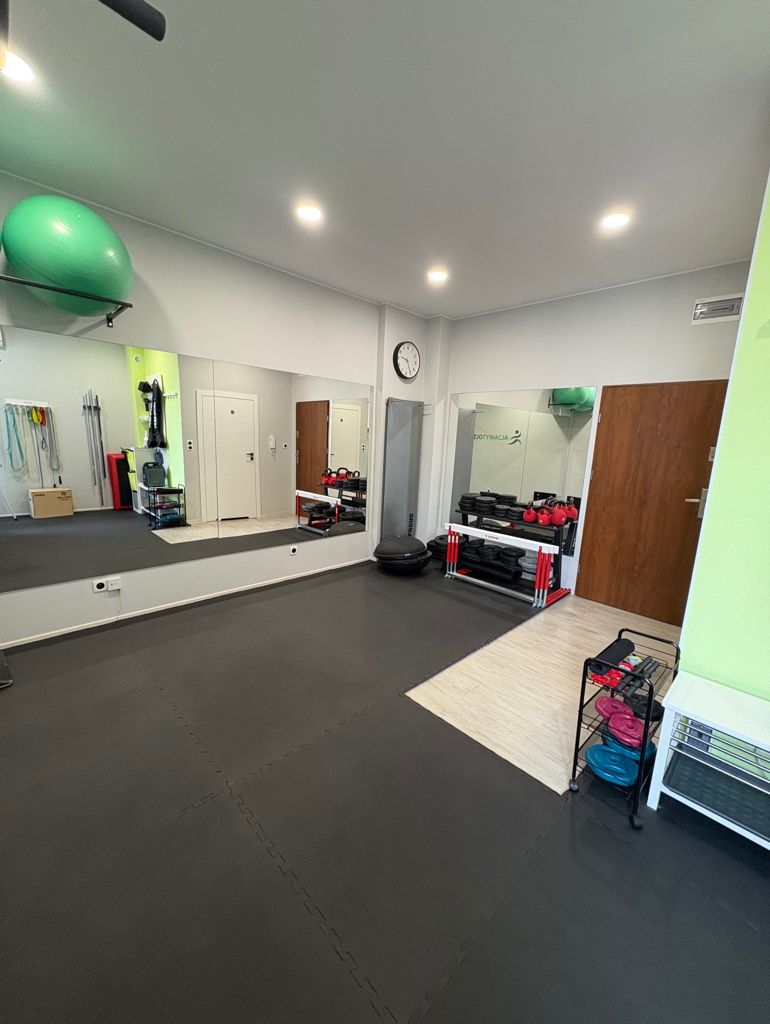 Sala do ćwiczeń z lustrami, wyposażona w sprzęt: piłka gimnastyczna, hantle, kettlebells. Podłoga wyłożona matą, widoczny zegar. Jasne, stonowane kolory.