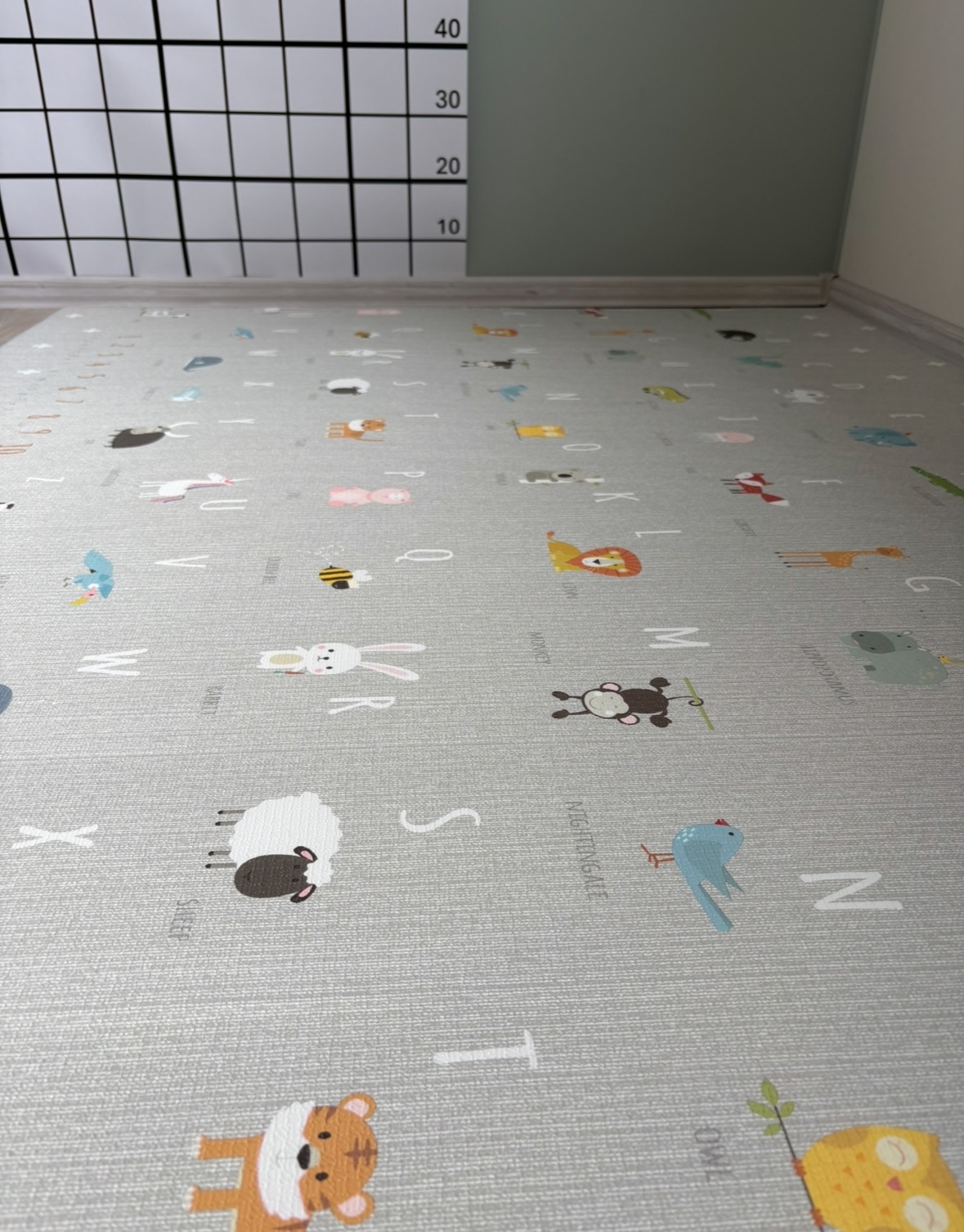 Szara mata edukacyjna dla dzieci z motywami zwierząt i liter alfabetu, ułożona na podłodze, widoczna z perspektywy podłogi, z fragmentem miarki wzrostu na ścianie.