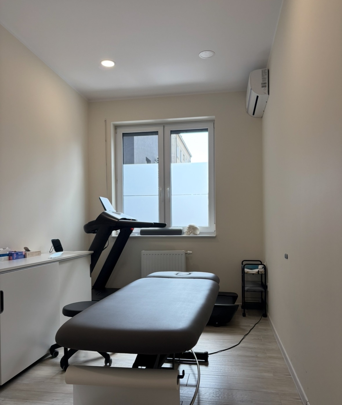 Jasne wnętrze gabinetu z kozetką, bieżnią i biurkiem. Na parapecie modelu czaszki. Minimalistyczny wystrój, stonowana kolorystyka, światło dzienne.