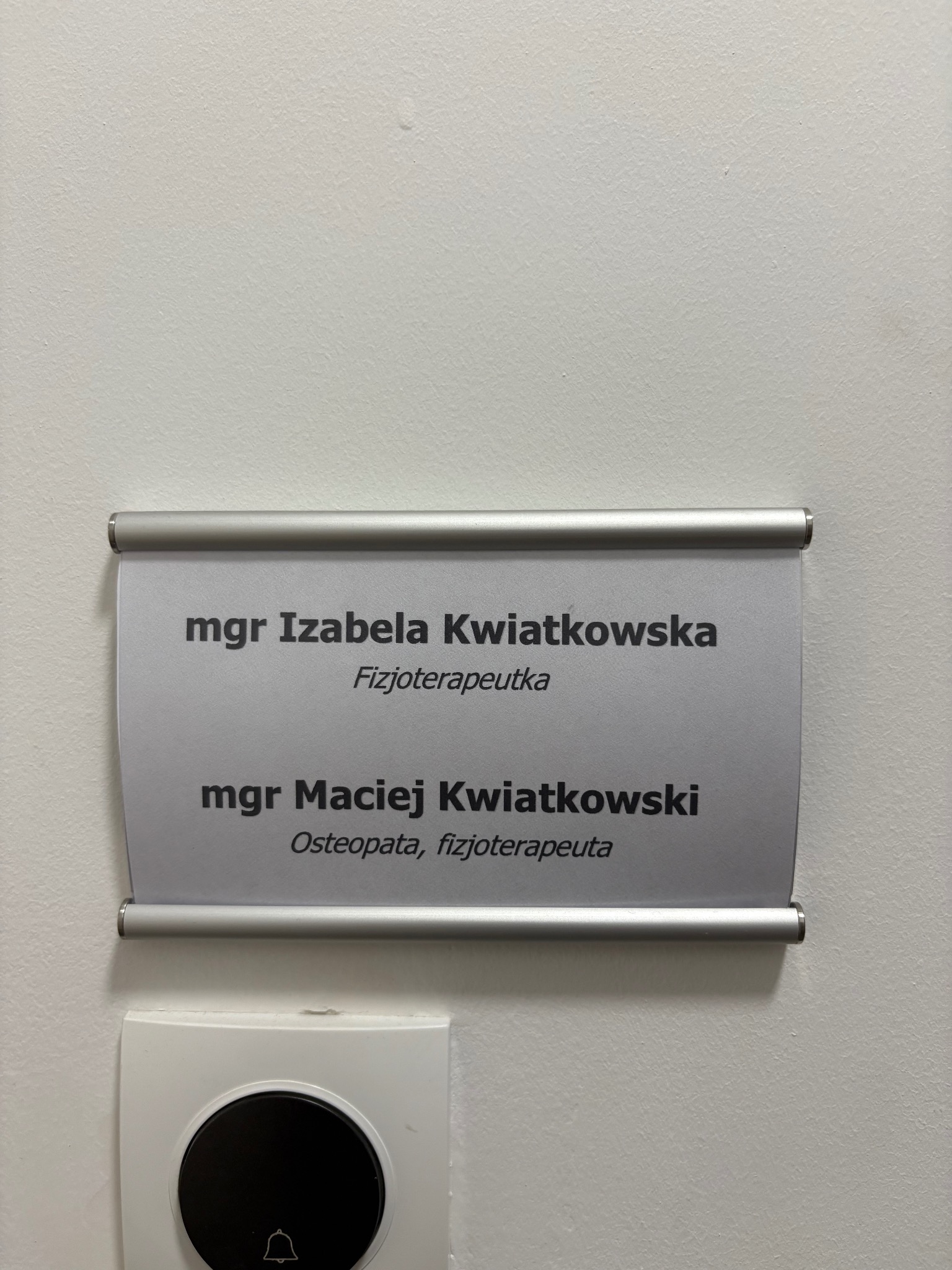 Tabliczka z nazwiskami mgr Izabeli Kwiatkowskiej, fizjoterapeutki oraz mgr Macieja Kwiatkowskiego, osteopaty i fizjoterapeuty, zamontowana na białej ścianie obok dzwonka.