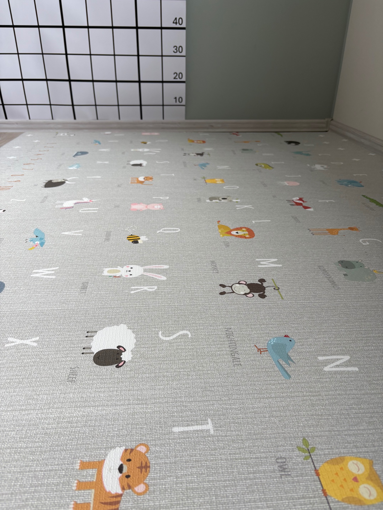 Szara mata edukacyjna z literami i zwierzętami na podłodze w Kaliszu, widok z niskiej perspektywy, z fragmentem ściany i miarką w tle.