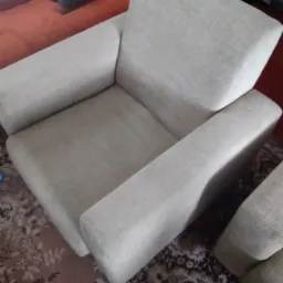 Jasny fotel tapicerowany o prostych kształtach, ustawiony na wzorzystym dywanie. Widoczny fragment sofy w tle.