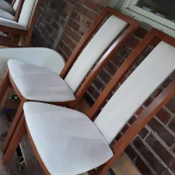 Krzesła z drewnianą ramą i jasną tapicerką, ustawione na palecie, z widoczną ceglaną ścianą w tle. Tapicerka krzeseł wydaje się być świeżo po renowacji lub w trakcie malowania.