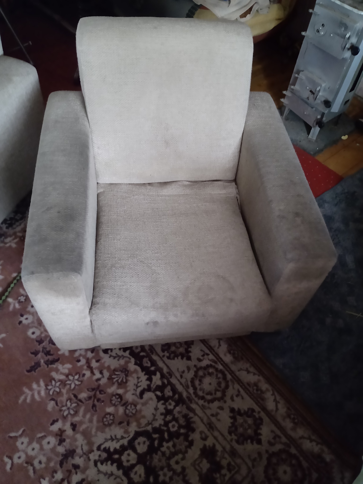 Beżowy fotel tapicerowany z widocznymi plamami i zabrudzeniami na siedzisku i podłokietnikach, stojący na wzorzystym dywanie, z piecem w tle.