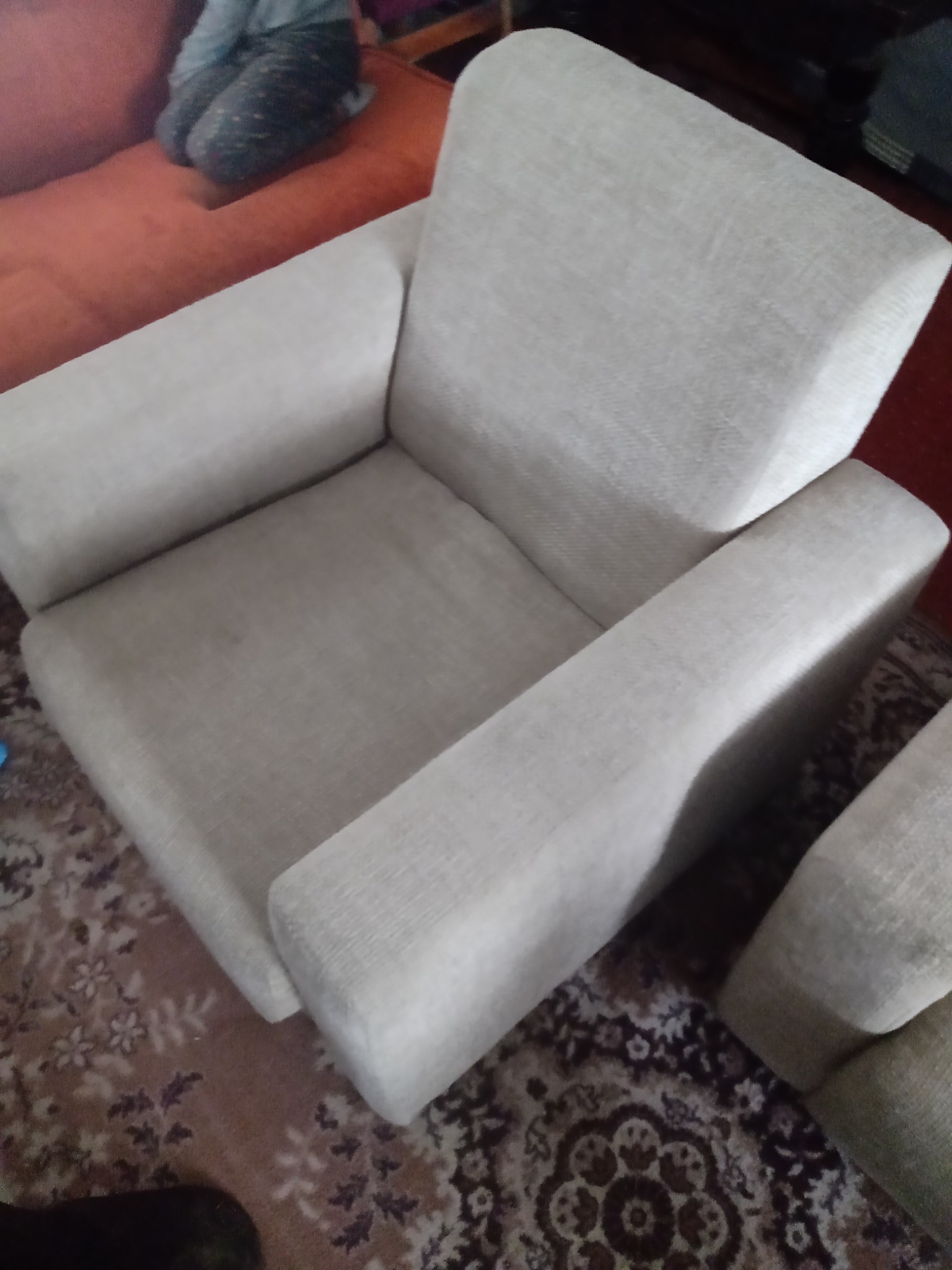 Jasny fotel tapicerowany o prostych kształtach, ustawiony na wzorzystym dywanie. Widoczny fragment sofy w tle.