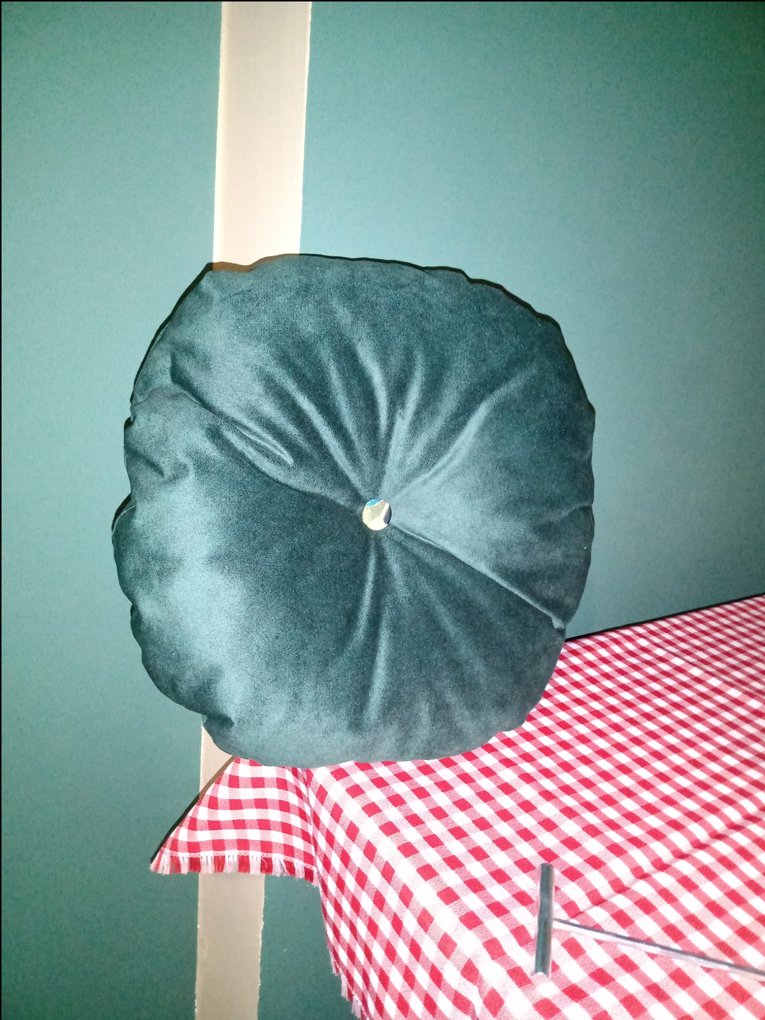 Okrągła, ciemnozielona poduszka z aksamitną fakturą i ozdobnym guzikiem na środku, leżąca na czerwono-białym obrusie w kratę, z widocznym narzędziem tapicerskim obok.