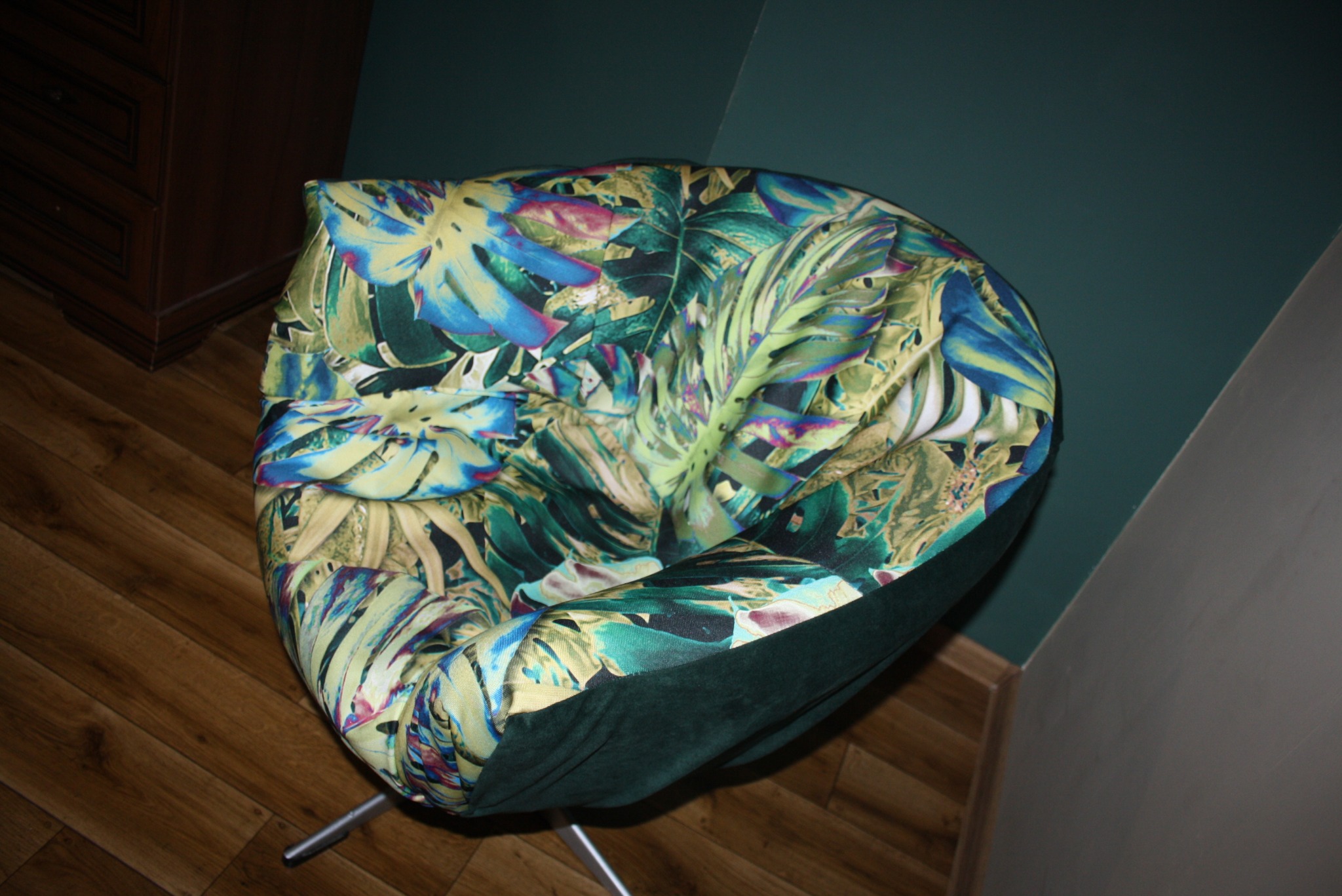 Krzesło tapicerowane w tropikalne liście na metalowej nodze, stojące na drewnianej podłodze przy ciemnej ścianie.