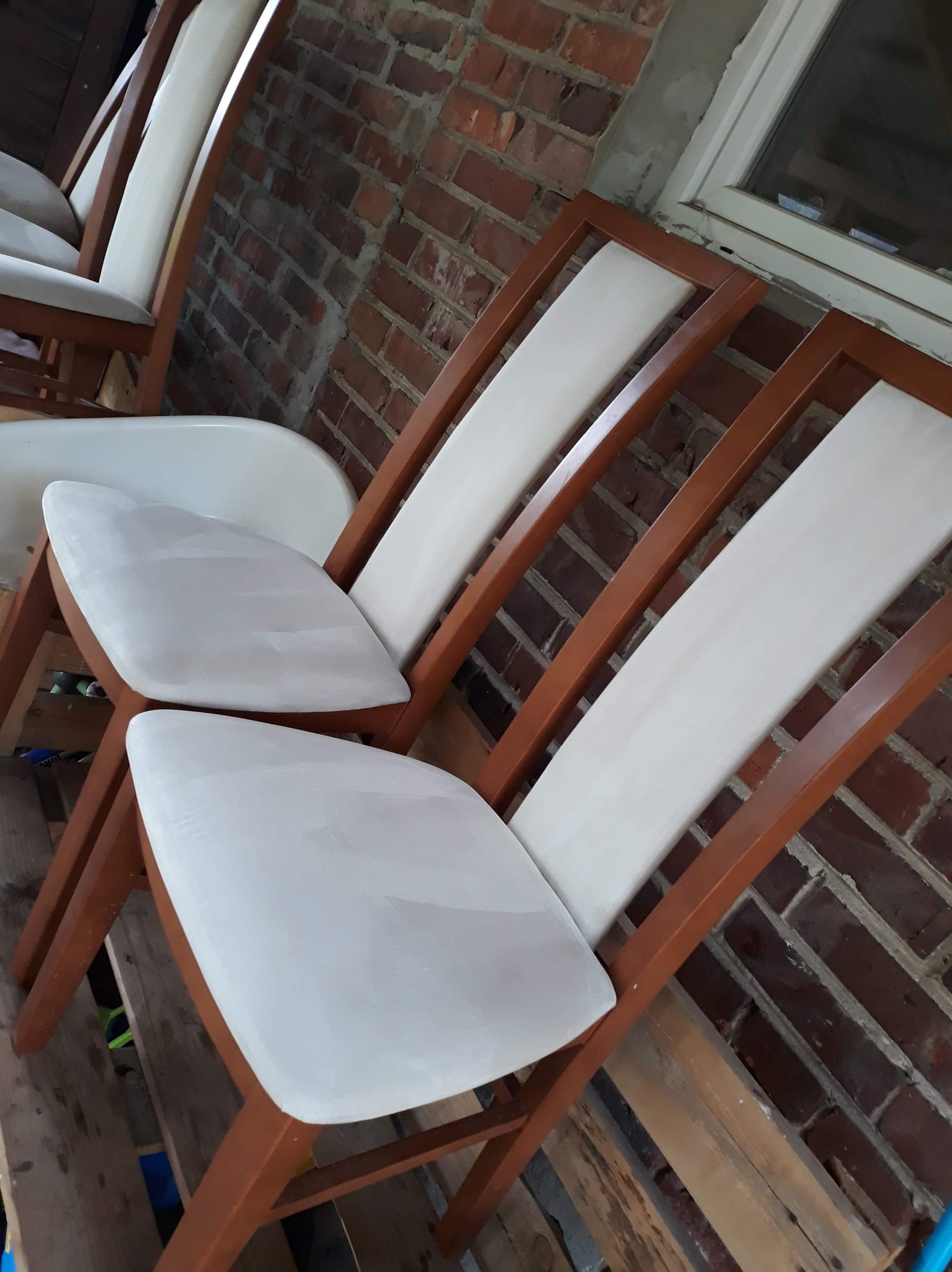 Krzesła z drewnianą ramą i jasną tapicerką, ustawione na palecie, z widoczną ceglaną ścianą w tle. Tapicerka krzeseł wydaje się być świeżo po renowacji lub w trakcie malowania.