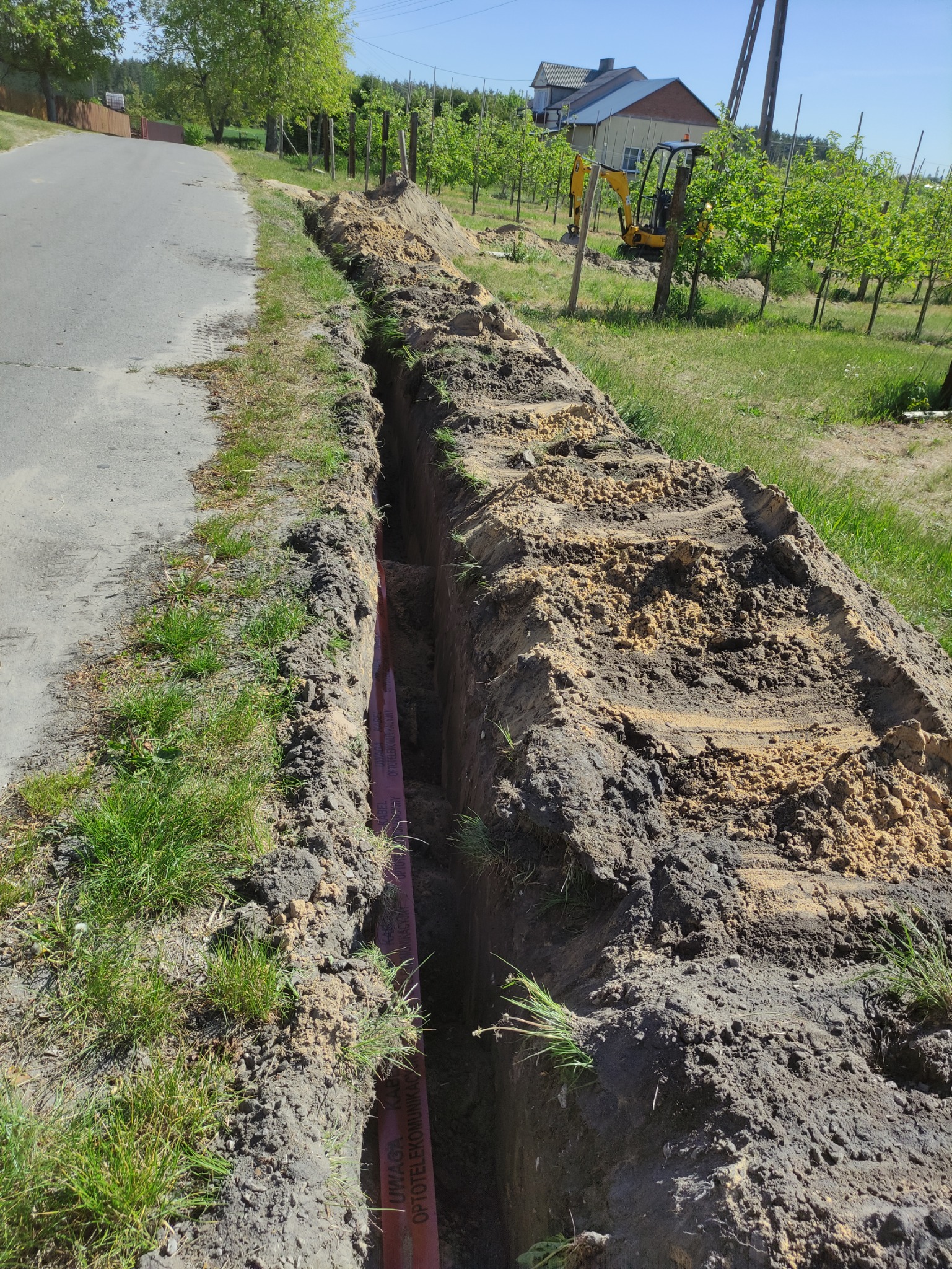 Wykopany wąski rów w ziemi wzdłuż drogi asfaltowej, z widoczną pomarańczową rurą kablową wewnątrz, niewielka koparka w tle.