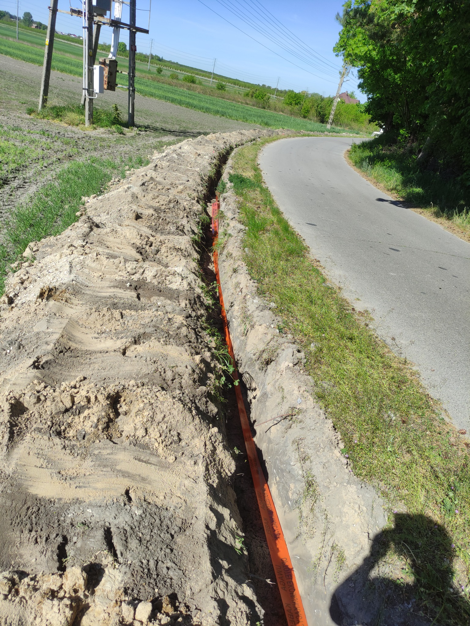 Wykopany wąski rów w ziemi wzdłuż drogi asfaltowej, z pomarańczową rurą instalacyjną wewnątrz, widoczny słup elektryczny i zielone pole w tle.