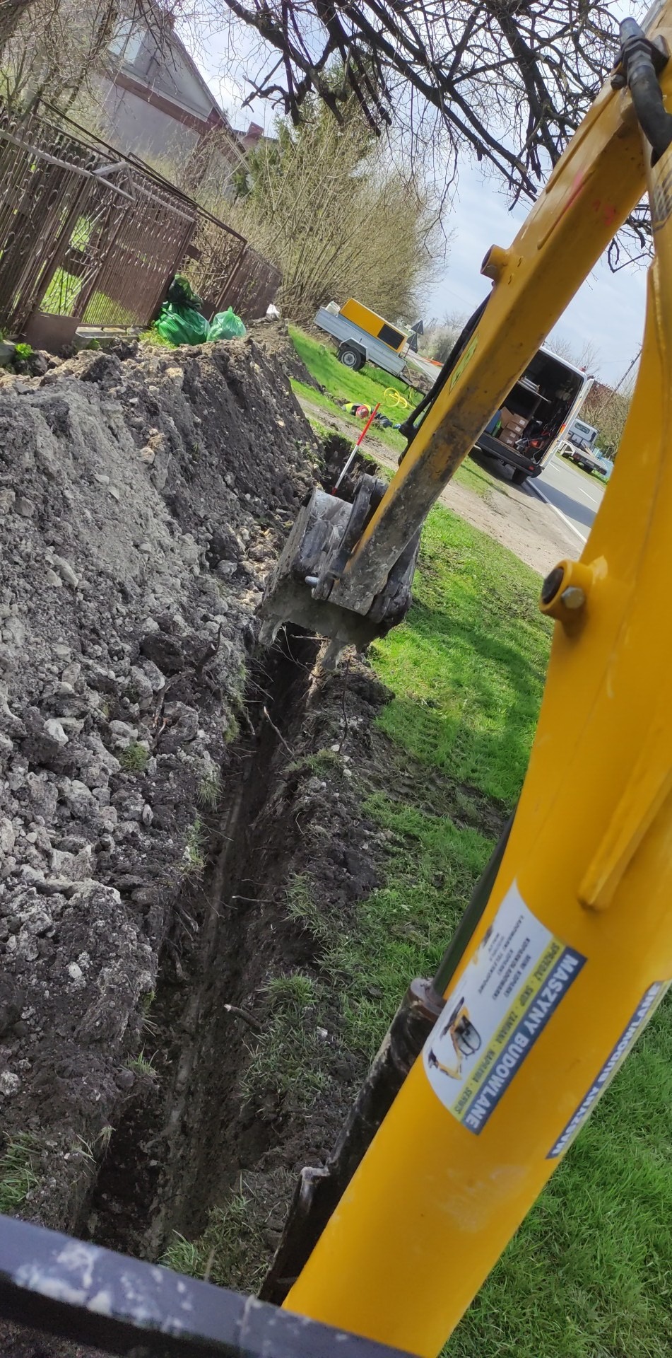 Żółta koparka w trakcie wykonywania wąskiego wykopu w ziemi, widoczny fragment łyżki koparki zanurzony w wykopie, w tle trawa, ogrodzenie i zaparkowane pojazdy.