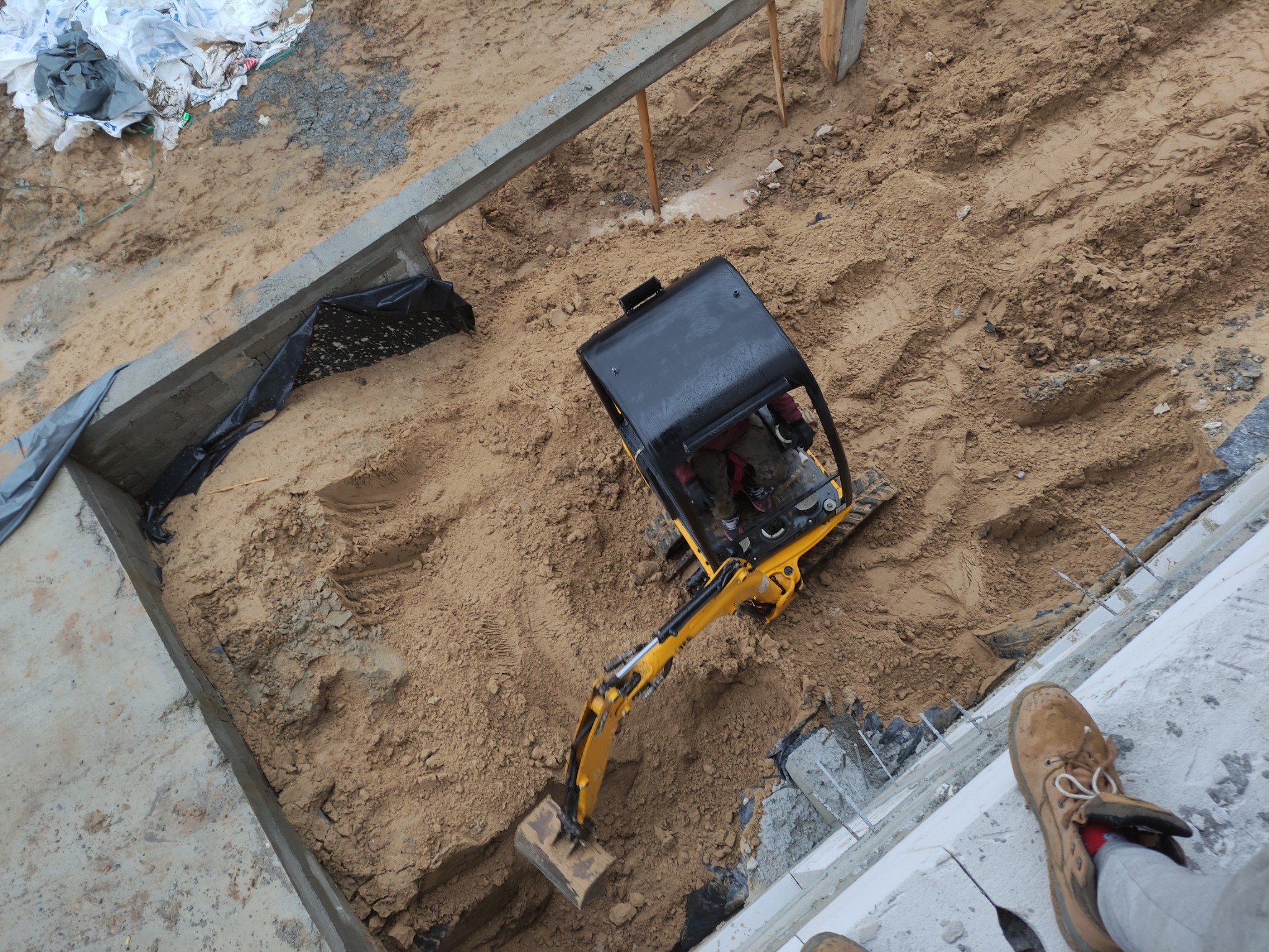 Żółta mini koparka w trakcie pracy przy wykopie pod fundamenty, widok z góry, obok widoczne zbrojenia i buty robocze.