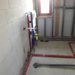 Surowy stan instalacji hydraulicznej i grzewczej w trakcie budowy, widoczne czerwone rury grzewcze z pomarańczowymi złączkami, szara rura kanalizacyjna, stelaż podtynkowy WC na tle ściany z białych...