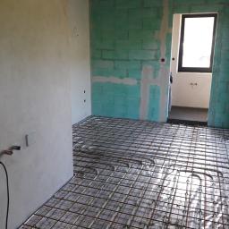 Instalacje - Wnętrze w trakcie instalacji ogrzewania podłogowego: rury grzewcze ułożone na folii, przytwierdzone do metalowej siatki, surowe ściany z widocznymi przyłączami elektrycznymi.
