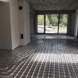 Wnętrze surowego budynku z widoczną instalacją ogrzewania podłogowego, rurki grzewcze ułożone w pętle na siatce zbrojeniowej, duże okno z widokiem na zieleń.