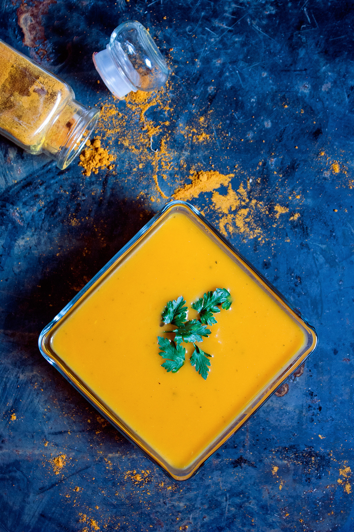 Kremowa zupa dyniowa w szklanym kwadratowym naczyniu, udekorowana świeżą natką pietruszki, obok rozsypana kurkuma i otwarta szklana buteleczka z przyprawą na ciemnym, teksturowanym tle.