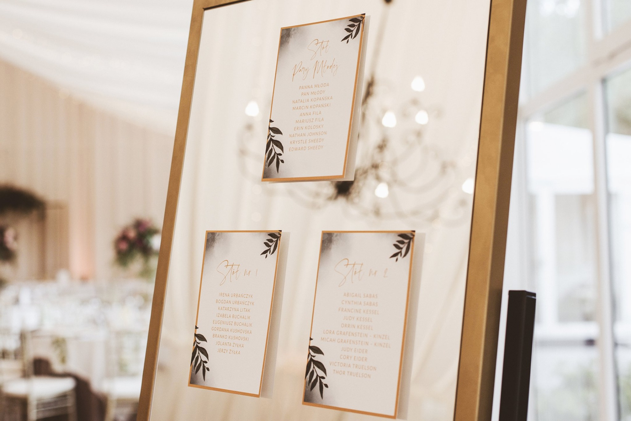 Tablica z listą gości weselnych w złotej ramie, z trzema kartami z imionami, ozdobionymi motywem liści i akwarelowymi plamami, ustawiona na tle sali z kwiatami i oknami.
