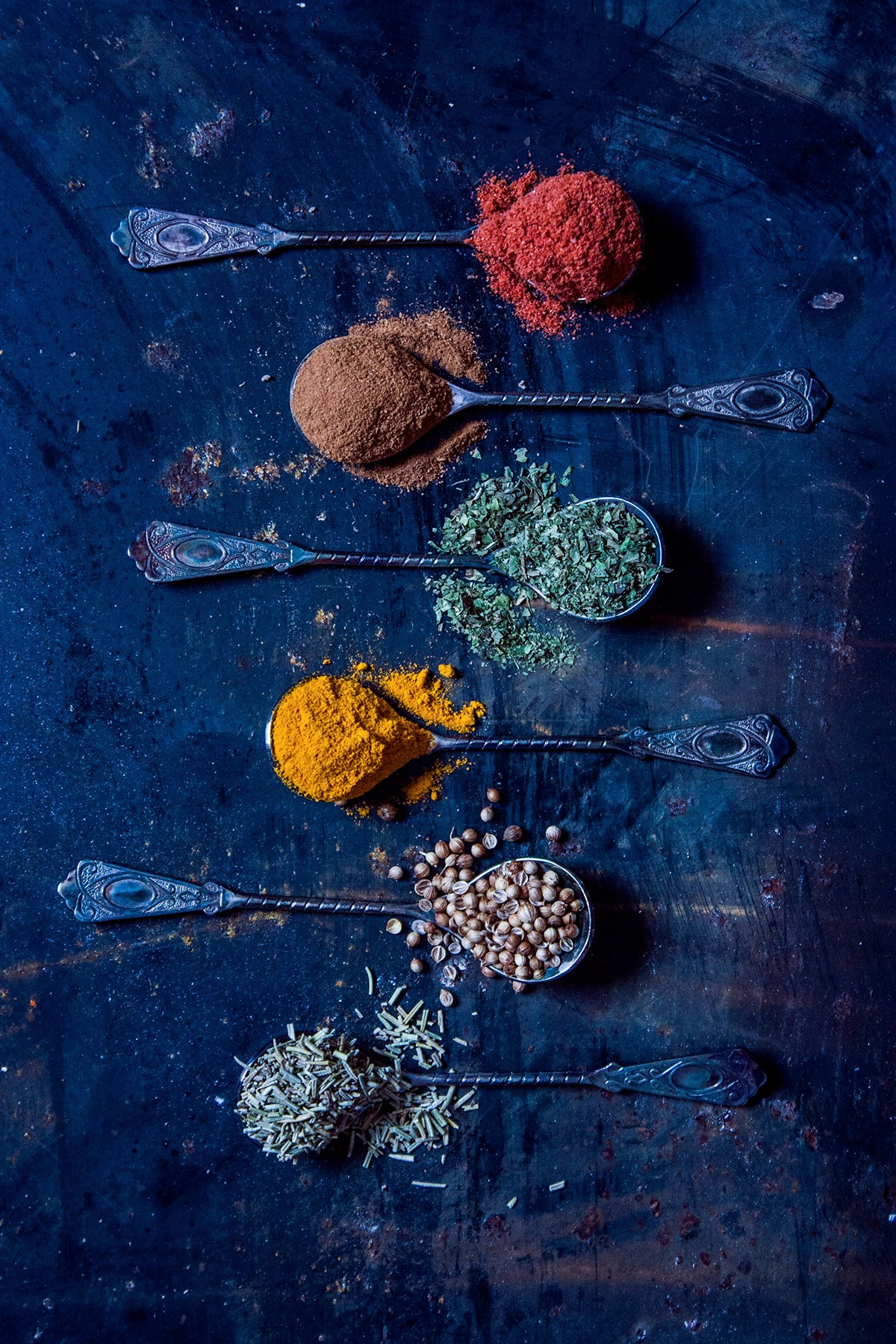 Sześć ozdobnych łyżeczek ułożonych pionowo, każda wypełniona inną przyprawą: papryka, cynamon, oregano, kurkuma, kolendra, rozmaryn, na ciemnym, metalicznym tle w Kobiór.