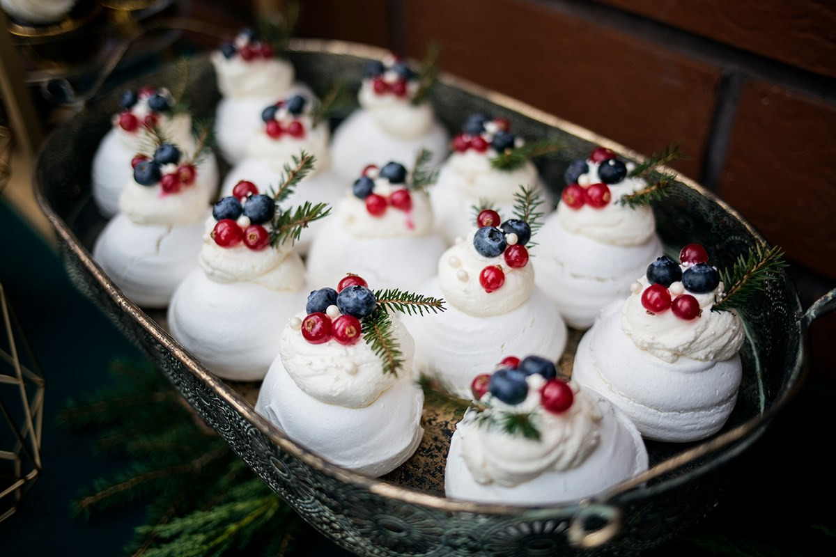 Mini bezy Pavlova udekorowane kremem, czerwonymi i niebieskimi jagodami oraz gałązkami świerku, ułożone na ozdobnej, srebrnej tacy.