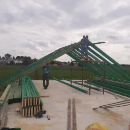 Montaż drewnianej konstrukcji dachu na budynku w Orchowie, widoczni dwaj mężczyźni pracujący na rusztowaniu, przygotowane elementy konstrukcyjne pomalowane na zielono.