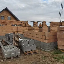 Budowa domu jednorodzinnego w stanie surowym otwartym: wzniesione ściany z czerwonej cegły ceramicznej, widoczne zbrojenie słupa, palety z bloczkami betonowymi na placu budowy, dom sąsiada w tle.