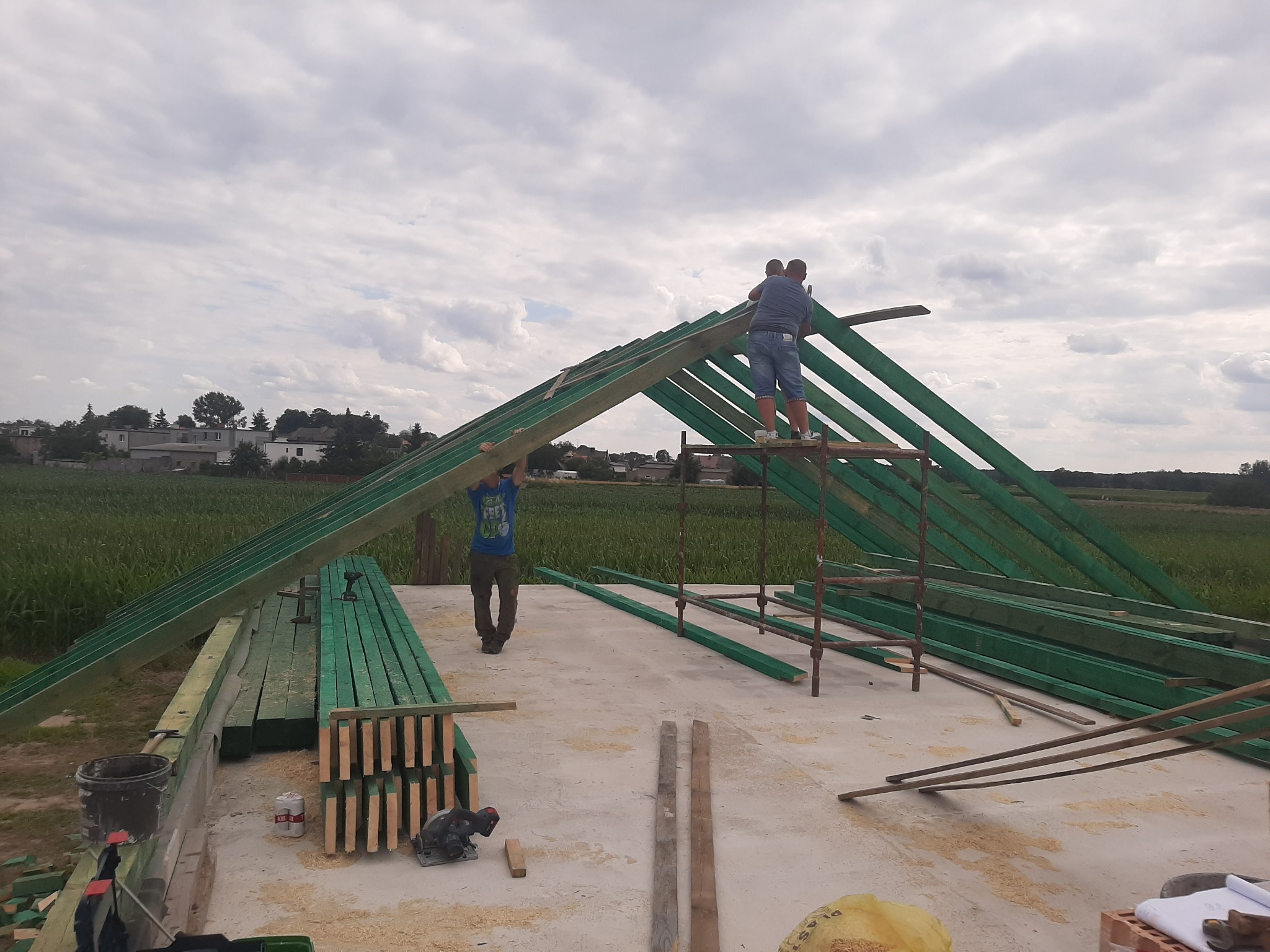 Montaż drewnianej konstrukcji dachu na budynku w Orchowie, widoczni dwaj mężczyźni pracujący na rusztowaniu, przygotowane elementy konstrukcyjne pomalowane na zielono.