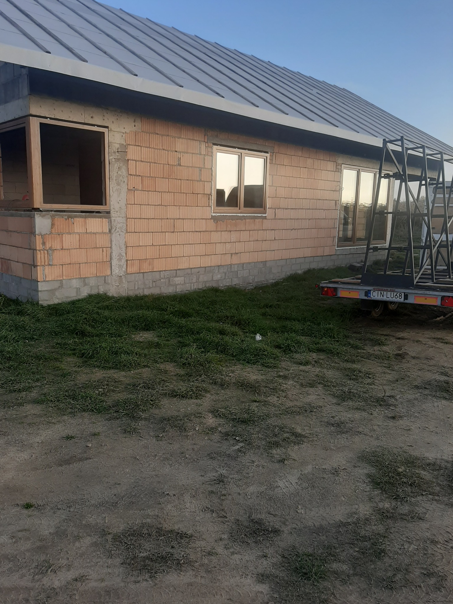 Surowa bryła domu w trakcie budowy z widocznym fragmentem dachu pokrytego szarą blachą, oknami z brązowymi ramami oraz przyczepą z metalową konstrukcją na tle nieba.