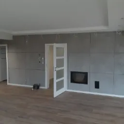 Surowe wykończenie salonu z panelami imitującymi beton, otwartymi drzwiami i kominkiem, widoczne przewody od oświetlenia sufitowego.