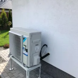 Zewnętrzna jednostka klimatyzacji Daikin zamontowana na metalowym stelażu przy białej ścianie budynku, widoczne przewody wchodzące w ścianę, kostka brukowa na ziemi.