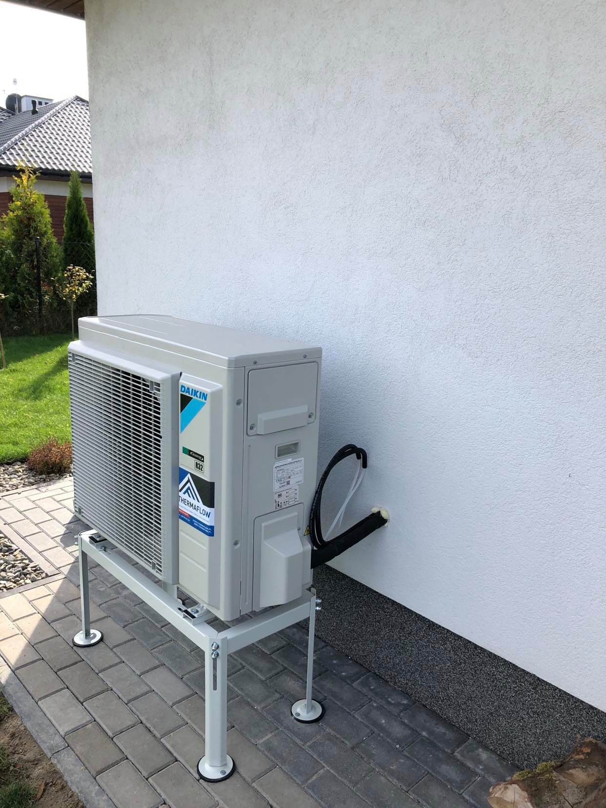 Zewnętrzna jednostka klimatyzacji Daikin zamontowana na metalowym stelażu przy białej ścianie budynku, widoczne przewody wchodzące w ścianę, kostka brukowa na ziemi.