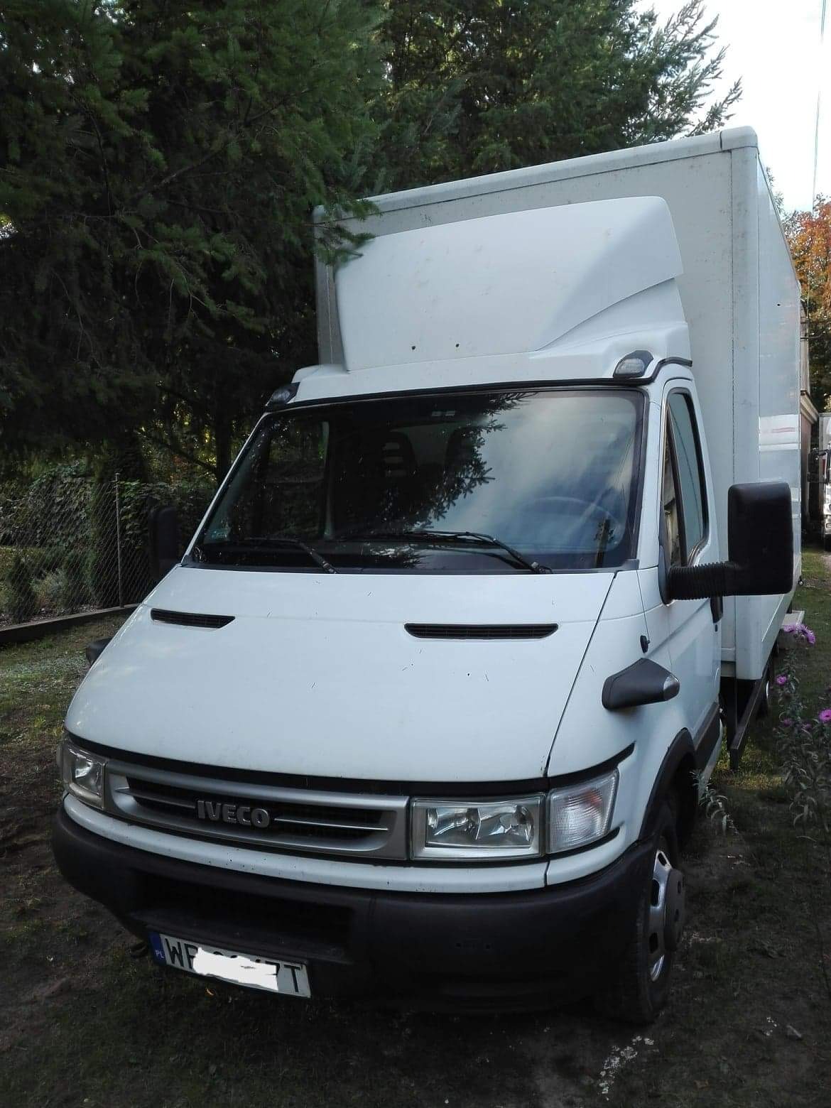 Biały samochód dostawczy Iveco z zabudową kontenerową, zaparkowany na trawie przed drzewami i ogrodzeniem z siatki.