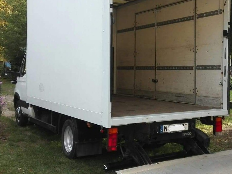 Biały samochód dostawczy z otwartymi tylnymi drzwiami, widoczne wnętrze pustej przestrzeni ładunkowej z szynami mocującymi na ścianach. Rampa załadunkowa opuszczona, trawa w tle.