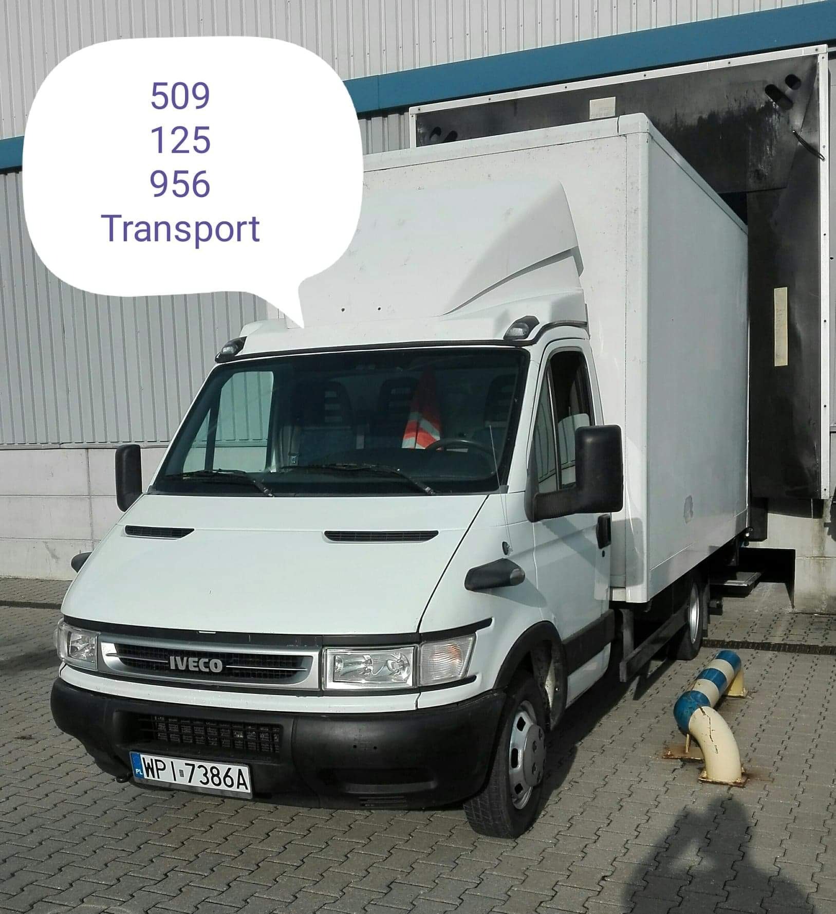 Biały samochód dostawczy Iveco z zabudową kontenerową, zaparkowany na kostce brukowej przed magazynem z rampą załadunkową. Widoczne niebiesko-białe zabezpieczenia rampy.