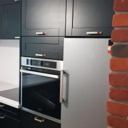 Ciemne, matowe szafki kuchenne z złotymi uchwytami, wbudowany piekarnik i srebrna lodówka, na tle ściana z czerwonej cegły.