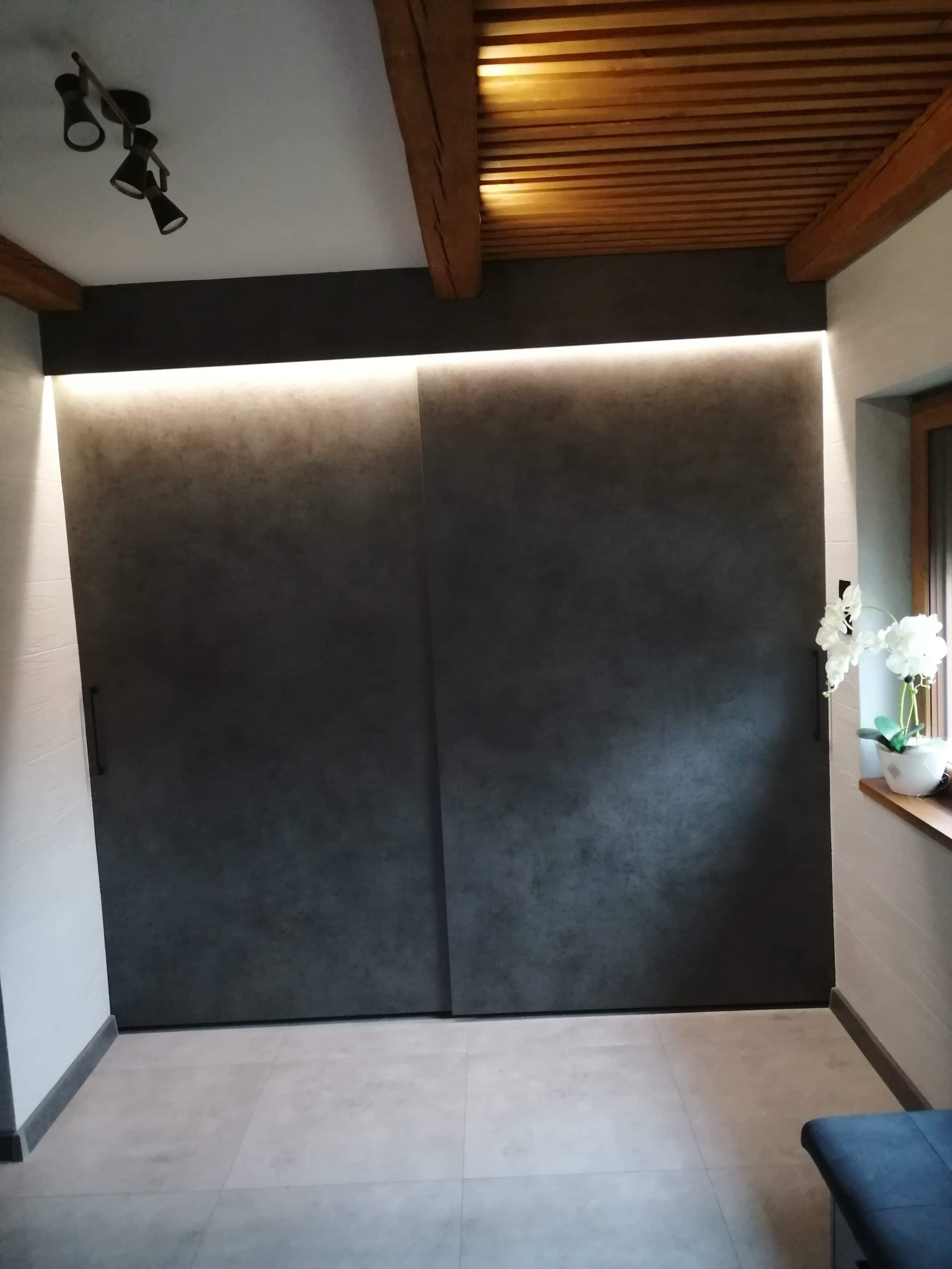 Nowoczesne, przesuwne drzwi w kolorze grafitowym z subtelnym oświetleniem LED nad nimi, drewniany sufit z belkami, białe ściany i podłoga w neutralnych odcieniach szarości, doniczka z białymi...