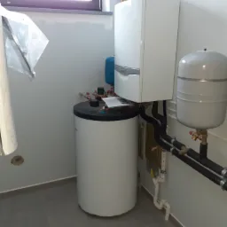 Biały zasobnik ciepłej wody użytkowej, wiszący kocioł gazowy z szarym paskiem i czarne rury w izolacji, zamontowane w pomieszczeniu z szarą podłogą i niedomalowaną ścianą.