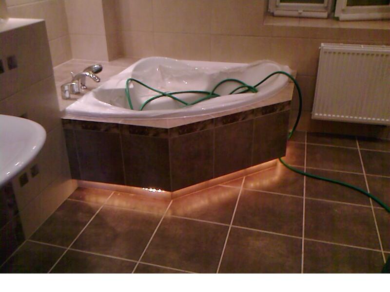 Nowoczesna narożna wanna z obudową wykończoną ciemnymi płytkami, podświetlona od dołu ciepłym światłem LED, na podłodze brązowe płytki, w tle grzejnik, wanna zabezpieczona folią ochronną.