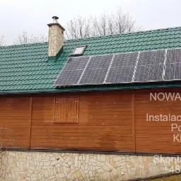 Drewniany budynek z zielonym dachem krytym blachodachówką, na którym zamontowane są panele fotowoltaiczne. Widoczny komin, okno dachowe oraz reklama firmy na ścianie budynku.