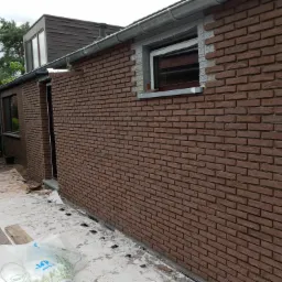 Brązowa ściana z cegły klinkierowej w trakcie układania, widoczne fragmenty ocieplenia wokół okna i folia ochronna na ziemi.