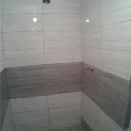 Wykończona łazienka z jasnymi i szarymi płytkami imitującymi drewno, z widocznym odpływem pod prysznicem i wystającymi elementami instalacji wodnej.