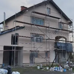 Dwupiętrowy dom w trakcie ocieplania elewacji, otoczony rusztowaniami, z widocznymi materiałami budowlanymi i błotnistym terenem wokół.