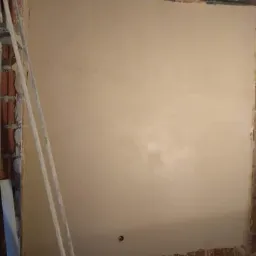 Świeżo nałożona, gładka warstwa szpachli na ścianie wewnętrznej, z widocznymi fragmentami cegieł i drewnianych elementów konstrukcyjnych w tle, oraz drabina oparta o ścianę po lewej stronie.
