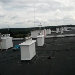 Widok z góry na płaski dach pokryty papą, z szeregiem białych obudów wentylacyjnych z metalowymi turbinami i instalacją odgromową, na tle zalesionego terenu i pochmurnego nieba.