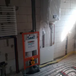 Surowa łazienka w trakcie remontu: widoczny grzejnik drabinkowy, stelaż podtynkowy JomoTech, rury instalacji podłogowej, piec gazowy zabezpieczony folią, surowe ściany z bloczków.