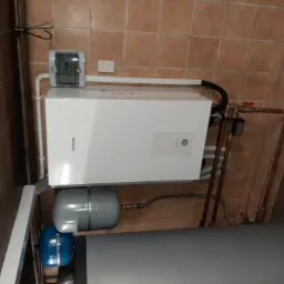 Biały piec grzewczy Panasonic zamontowany na ścianie z brązowych płytek, z widocznymi rurami miedzianymi i elementami instalacji, w pomieszczeniu technicznym.